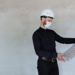 Architect in safety helmet and mask reviewing building plans indoors.