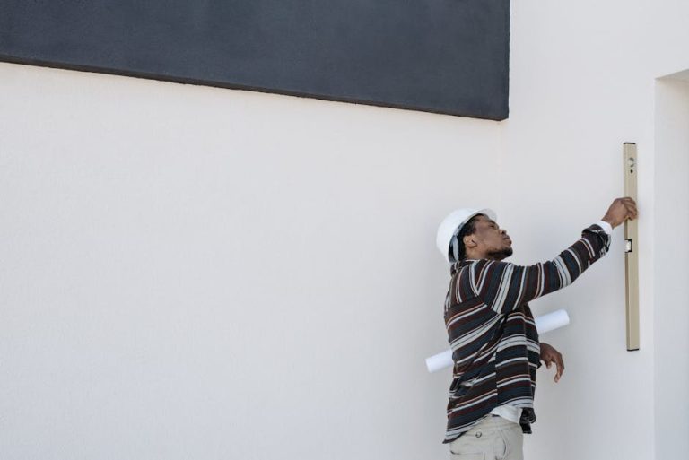 An architect wearing a hard hat checks wall alignment with a level tool outdoors.