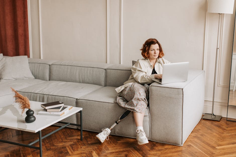 A woman with a prosthetic leg works remotely on a laptop in a cozy living room setting.