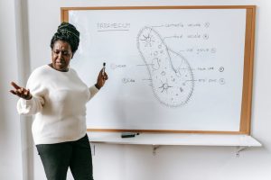 Expressive African American female teacher in casual clothes standing near whiteboard and explaining scheme during biology lesson in classroom