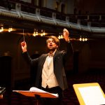 Bio-Inspired Edge Orchestration for Distributed Systems Orchestra conductor passionately leading a performance in an elegant concert hall.