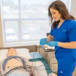 Medical student in blue scrubs practicing procedures on a patient training manikin in a clinical setting.