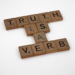 Scrabble tiles arranged on a white background spelling 'Truth is a Verb' for creative representation.