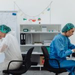 Two scientists in a lab, one examining a microscope, the other taking notes.