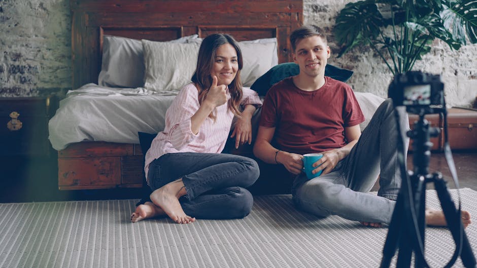 Casual couple sitting on bedroom floor, smiling at camera setup.