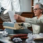 Elderly man with glasses using a vintage typewriter, surrounded by books and papers in a study.