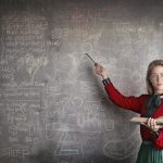 Serious female teacher wearing old fashioned dress and eyeglasses standing with book while pointing at chalkboard with schemes and looking at camera