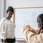 Attentive African American woman standing near whiteboard while explaining scheme to schoolgirl during lesson