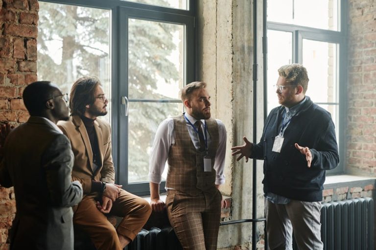 Four diverse professionals engaged in a strategic discussion near large windows in an industrial office setting.