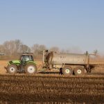 A tractor with a tanker spreading fertilizer over a barren farmland under clear skies.