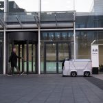 Few-Shot ISRU: Autonomous Material Synthesis for Extremes A modern office setting featuring a man and an autonomous delivery robot outside a glass-covered building.