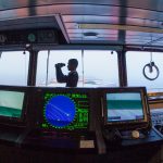 Silhouette of a navigator using binoculars on a cargo ship's bridge, surrounded by navigation equipment.