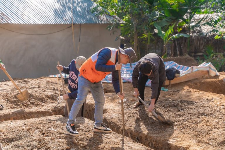 Men working at a construction site, digging foundation ditches with shovels in a rural area, outdoors.