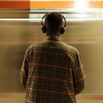 A man wearing headphones stands at a subway station, observing a passing train.