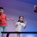 Low-Latency Embodied Intelligence: Beyond the Screen Two children joyfully dance with a robot in a colorful indoor setting.