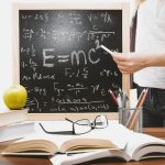 Woman writing physics equations on a blackboard with books and an apple on the desk.