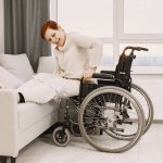 Optimizing Hospital at Home Supply Chains for Distribution Shift A woman with dyed short hair smiles while transitioning from a wheelchair to a couch.