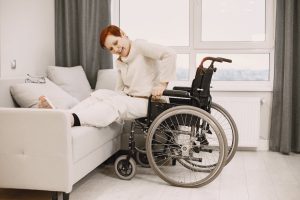 A woman with dyed short hair smiles while transitioning from a wheelchair to a couch.