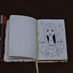 Outline Artistic sketch of a woman in an open notebook with pens on a dark surface.