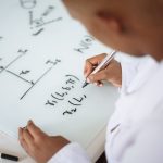 From above anonymous ethnic student wearing uniform and solving problem in chemistry while writing formula on white table in lab