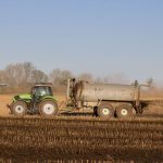 A tractor with a tanker spreading fertilizer over a barren farmland under clear skies.