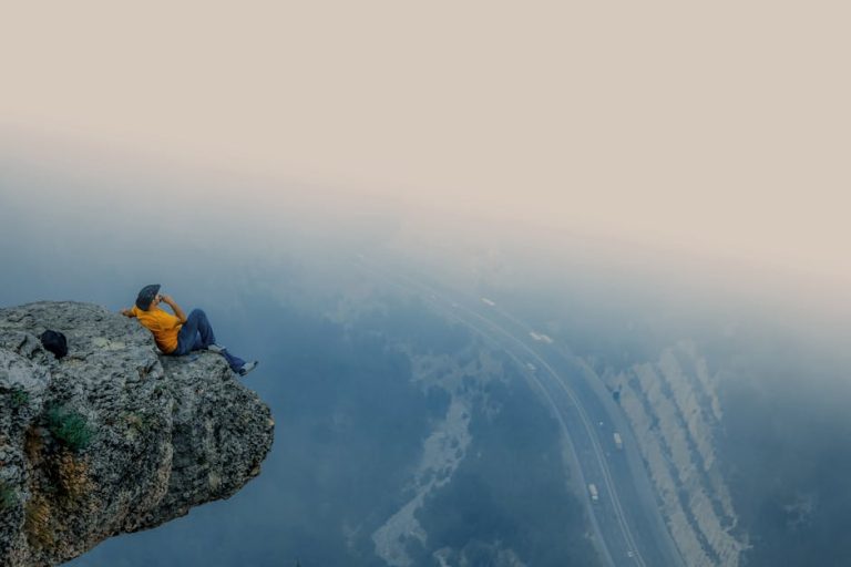 A man in an orange shirt sits on a rocky cliff edge overlooking a foggy road in Mersin, Turkey.