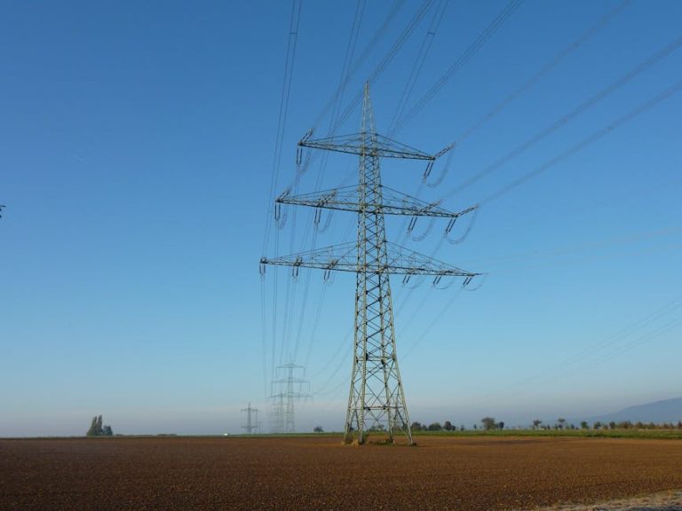 High voltage power lines on a tower standing in an open field against a clear blue sky.
