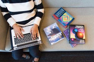 Woman using laptop on sofa, surrounded by programming books, learning coding.