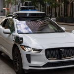 Self-Evolving TinyML: The Future of Autonomous Edge AI A white autonomous vehicle navigating a city street, reflecting urban architecture in daylight.