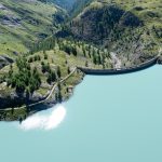 Sim2Real Climate Adaptation Compilers for Infrastructure A stunning aerial view of the dam and turquoise lake in Korutany, with lush green landscapes and clear blue skies.
