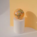 A globe rests on a white cylinder against a pastel yellow backdrop in a minimalist studio setting.