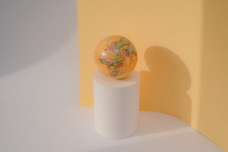 A globe rests on a white cylinder against a pastel yellow backdrop in a minimalist studio setting.