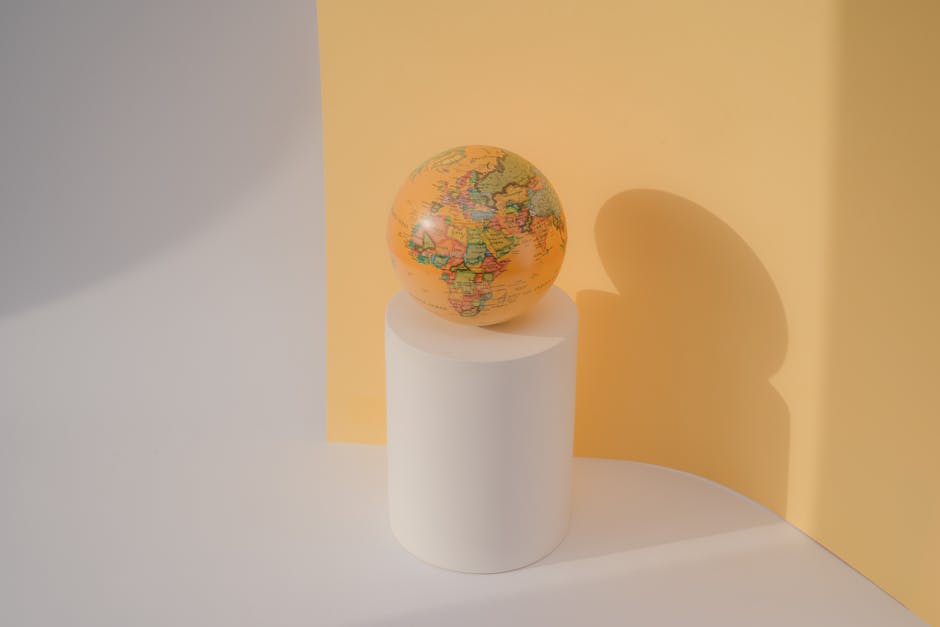 A globe rests on a white cylinder against a pastel yellow backdrop in a minimalist studio setting.