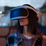 <h1>Simulation-To-Reality Topological Computing Standards for Distributed Ledgers</h1> Young woman exploring virtual reality with VR headset indoors, surrounded by colorful lights.
