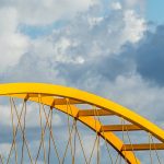 A close-up view of a yellow steel bridge arch contrasting with a dramatic cloudscape.