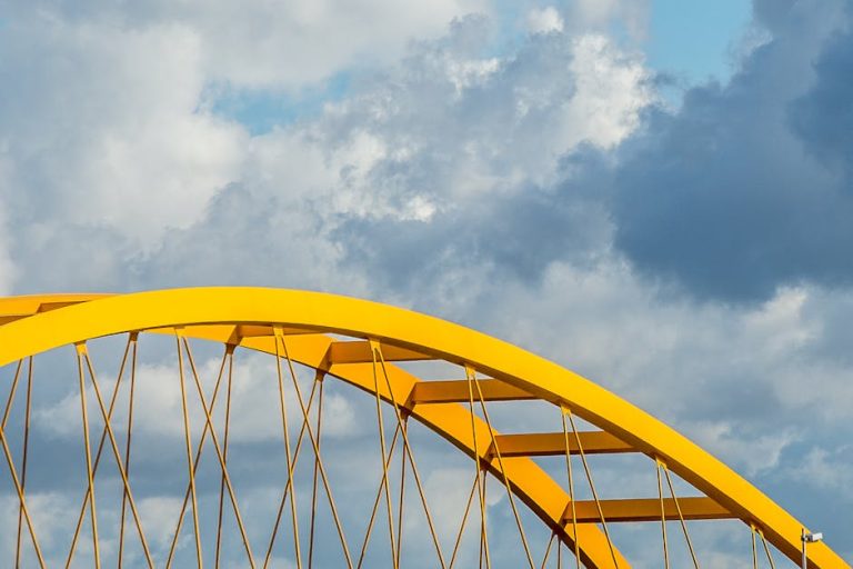 A close-up view of a yellow steel bridge arch contrasting with a dramatic cloudscape.