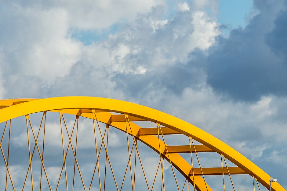 A close-up view of a yellow steel bridge arch contrasting with a dramatic cloudscape.