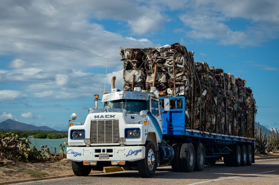 A Mack truck loaded with scrap metal travels on a rural road under a cloudy sky.