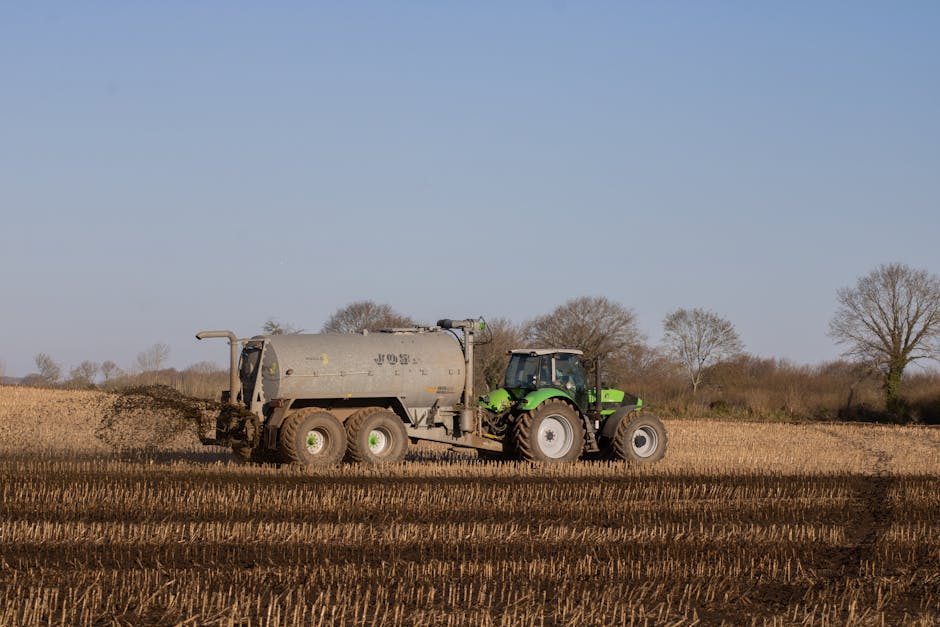 A green tractor spreading fertilizer on a brown, freshly plowed farmland under a clear sky.
