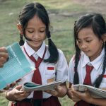 Systemic Literacy: Redesigning Education for Environmental Strategy Two young girls in school uniforms reading outdoors with a book in hand, captured in natural daylight.