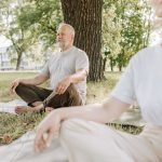Elderly man and woman meditating outdoors on yoga mats in a peaceful park setting.