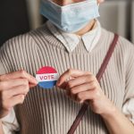 A person wearing a mask holds a voting badge, symbolizing participation in the election.