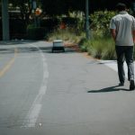 A man walks down a sidewalk beside a small delivery robot on a sunny day.