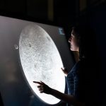 A woman interacts with a touchscreen showing the moon's surface, exploring lunar details.