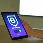 The Blockchain Mandate: Architecting Economic Systems for Scale A hand holding a tablet with blockchain logo on screen, showcasing digital technology.