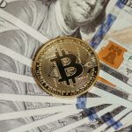 The Consciousness Shift: Why Economics Is Becoming Internal A Bitcoin coin placed on a stack of hundred dollar bills, symbolizing cryptocurrency and traditional wealth.