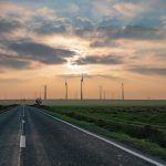 The Economic Edge of Renewable Energy Strategy A picturesque rural road leading to wind turbines under a vibrant sunset in Romania, emphasizing renewable energy.