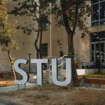 Modern building entrance with large STU letters surrounded by greenery and urban design.