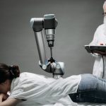A woman receives a robotic massage as a scientist monitors, showcasing modern technology.