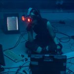 <h1>The Future of Bio-Computing: Building Low-Latency Programmable Biology Interfaces</h1> A woman in a dark room wearing a VR headset surrounded by retro technology.